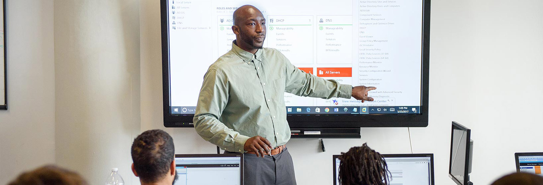 Instructor teaching a classroom of learners, pointing at a large screen displaying a software dashboard.