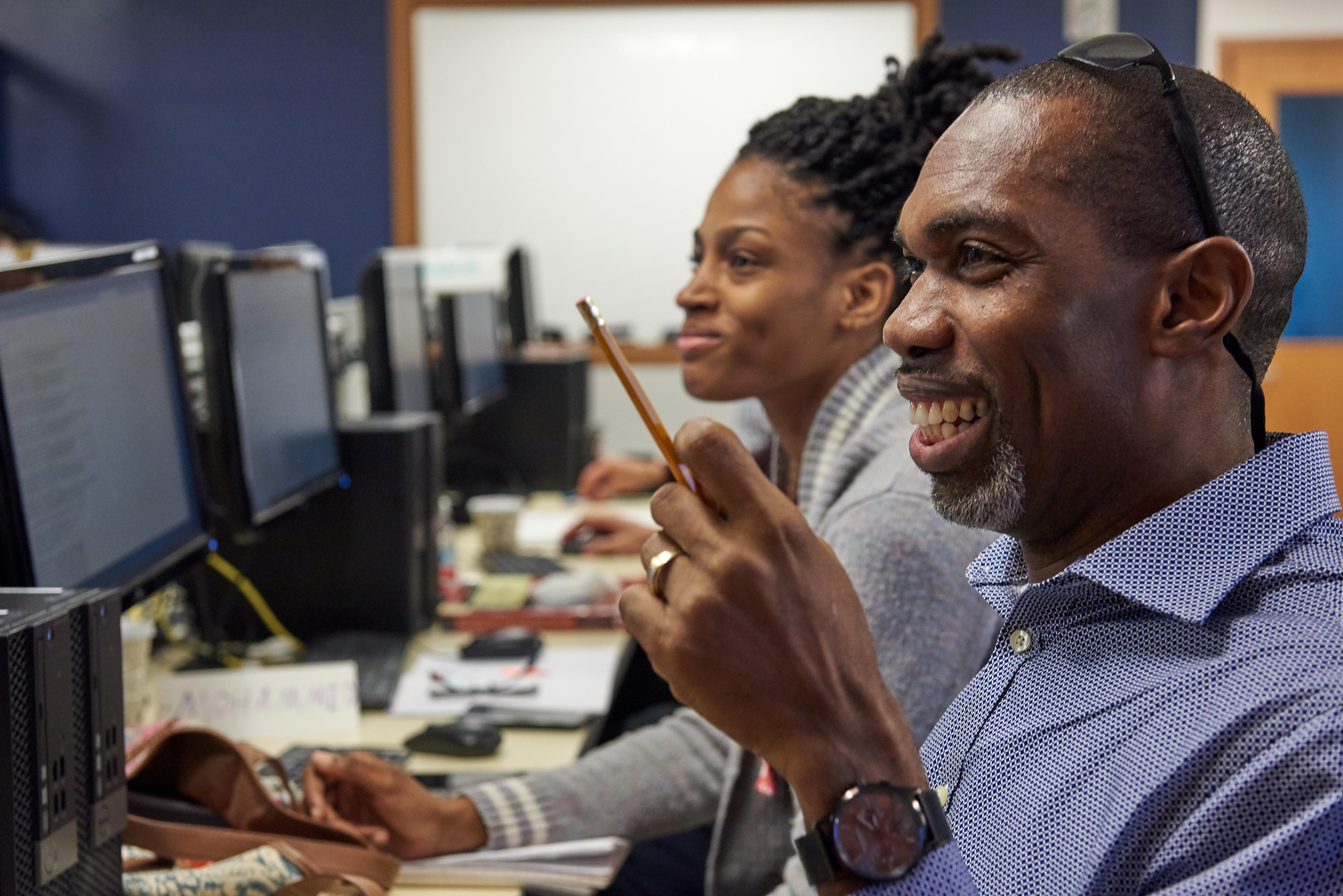 Two adult learners in a computer lab smiling while working at desktop computers.