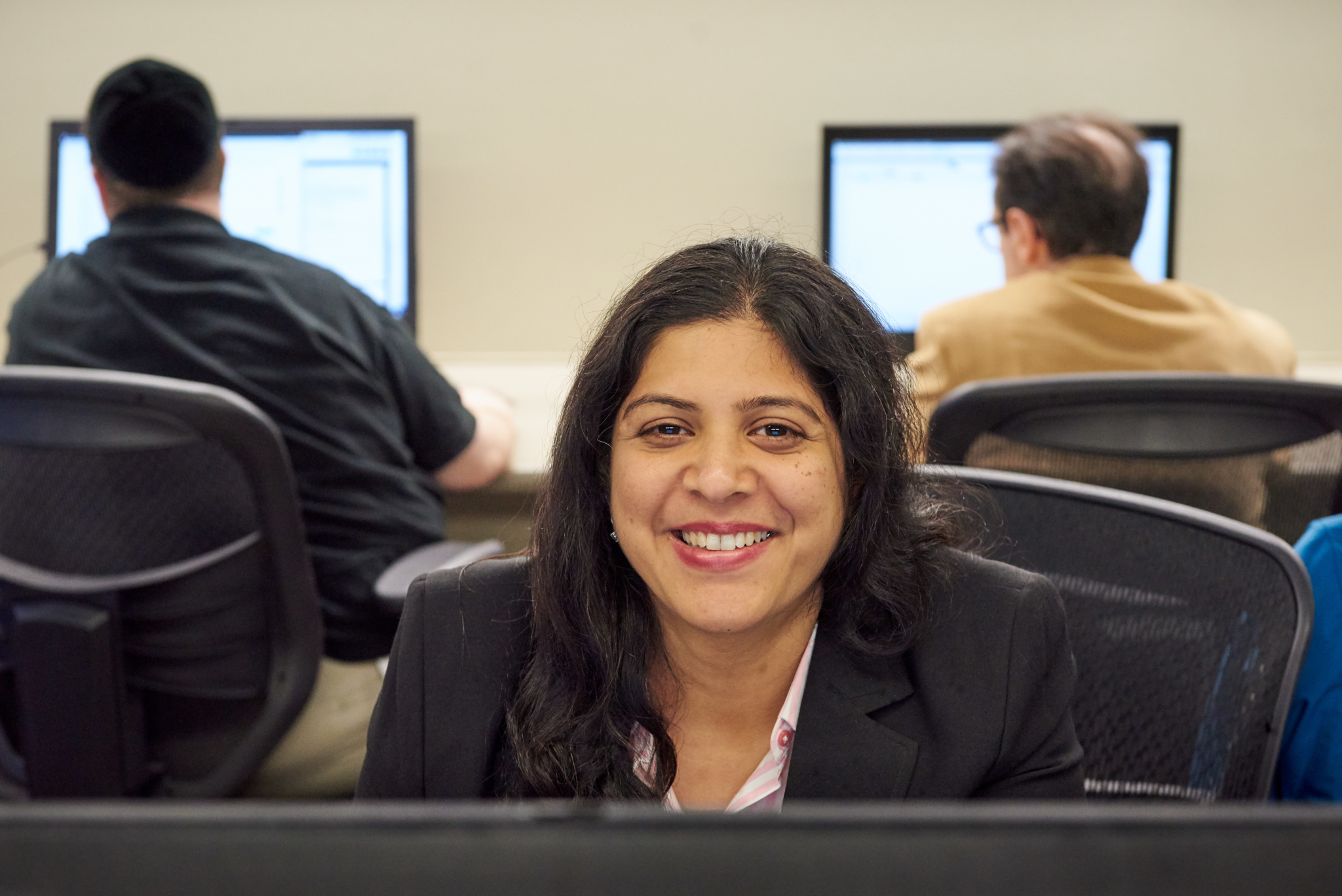 Woman smiling at camera while seated at a computer in a classroom with two other students working at monitors behind her.