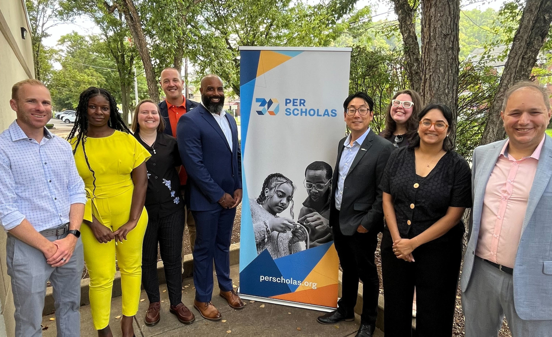 Group of Per Scholas staff and supporters standing outside around a Per Scholas banner, smiling for a photo.