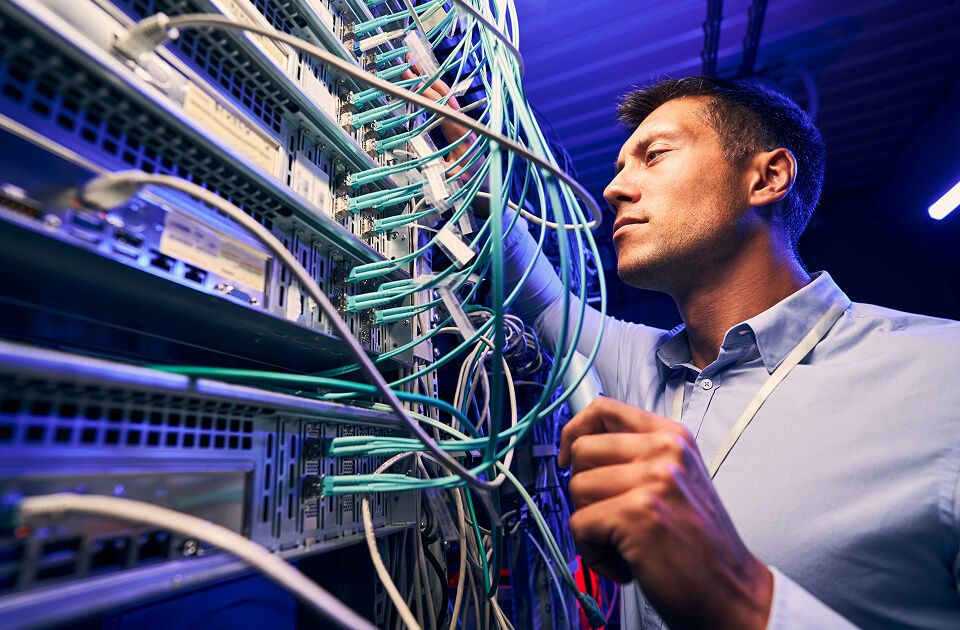 Man in a light blue shirt organizing network cables on a server rack, illuminated by blue lighting in a data center.