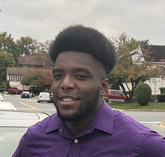 Raekwon Frazier smiling, while outside on a suburban street, wearing a purple button-down shirt.