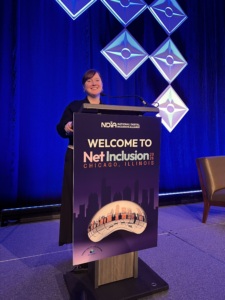 Woman standing at a podium speaking at a conference stage with a "Welcome to Net Inclusion Chicago, Illinois" sign.