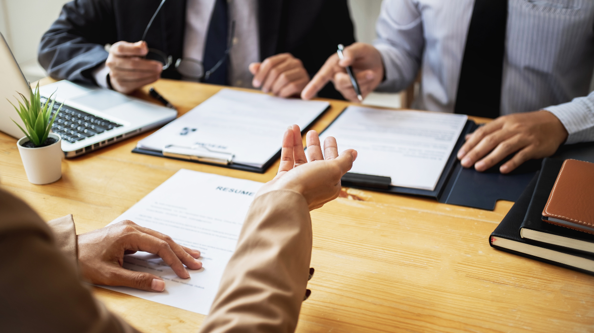 Candidate meeting with interviewers across a table, discussing a resume and pointing at documents.