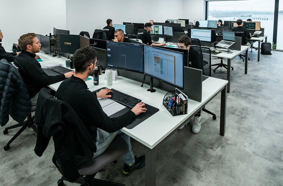 Several people focused on computer screens at rows of desks in a bright office space.