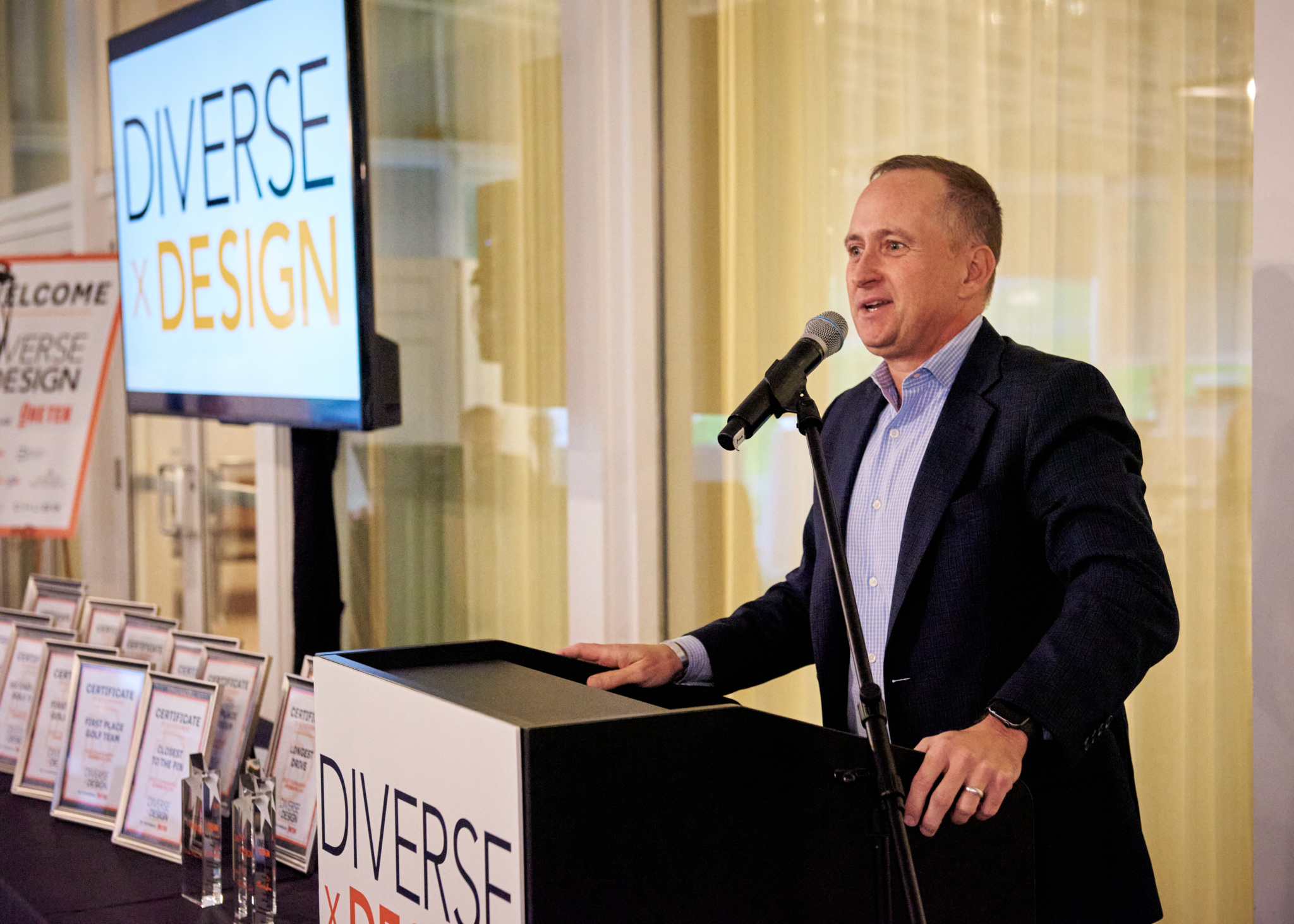 Man speaking at a podium with a microphone during a "DIVERSE x DESIGN" event; framed certificates and a screen showing the event name are visible.