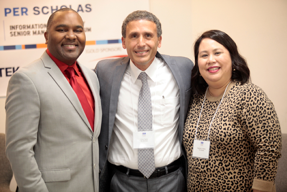 Three smiling professionals wearing conference name badges stand together in front of a Per Scholas event sign.