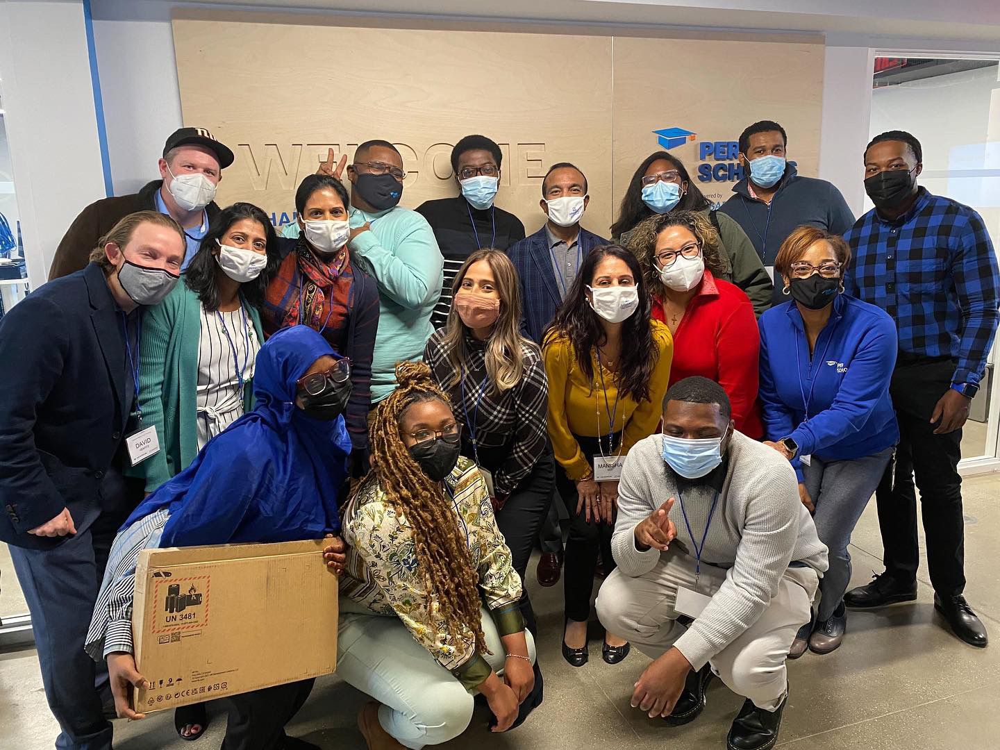 Group of students and staff wearing masks posing together in a classroom lobby, some kneeling and one holding a boxed laptop.