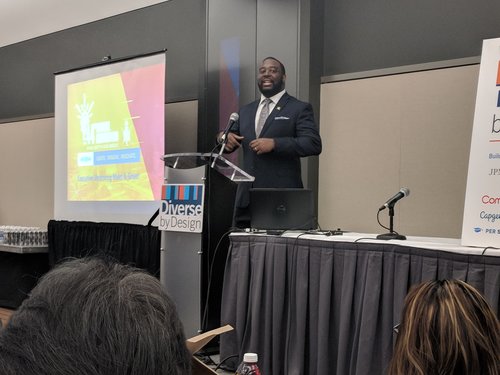 Man standing at a podium speaking to an audience at a conference, with a presentation slide and a "Diverse by Design" sign visible.