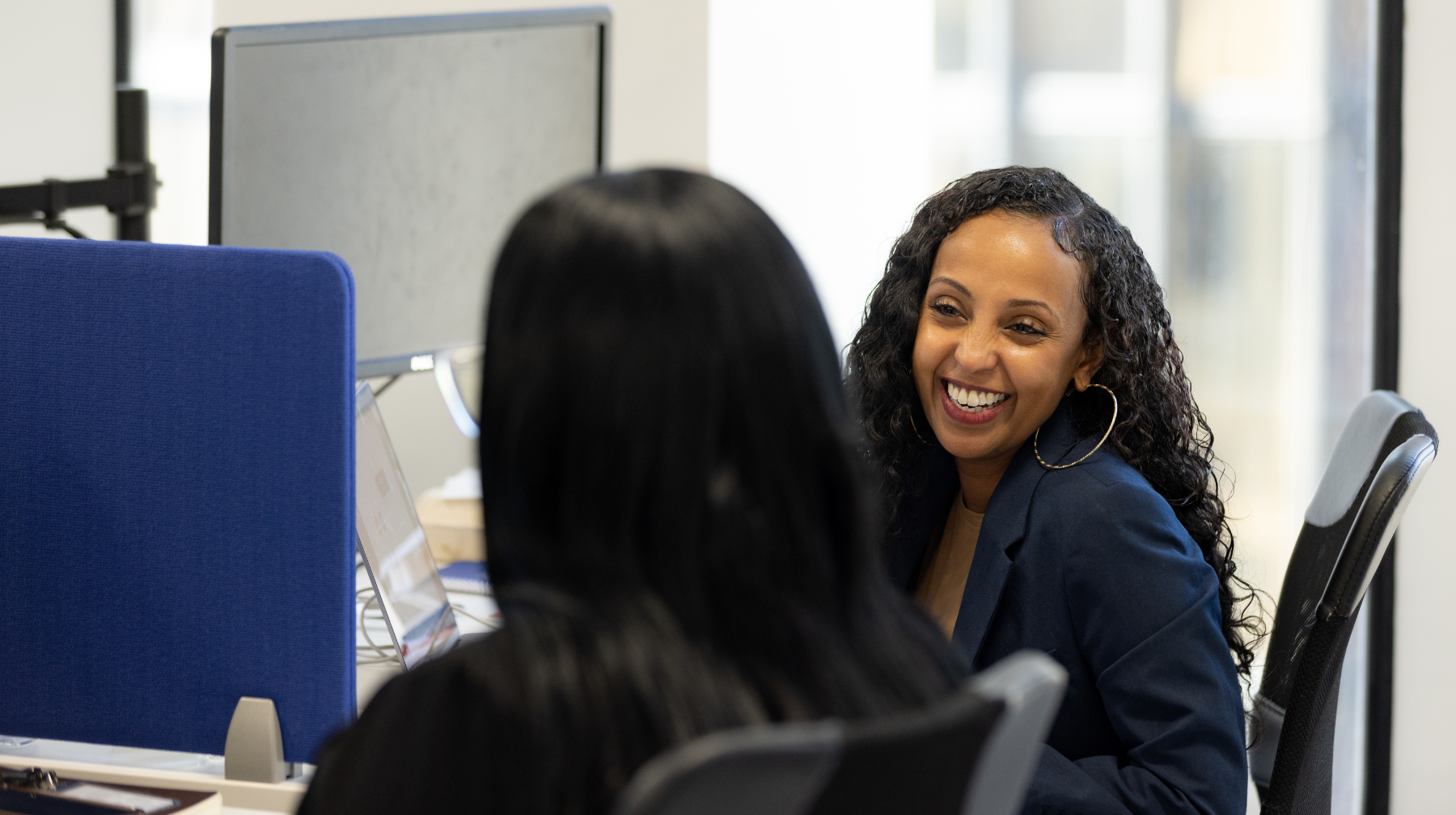 Two women smiling and talking at adjacent desks in an office setting.