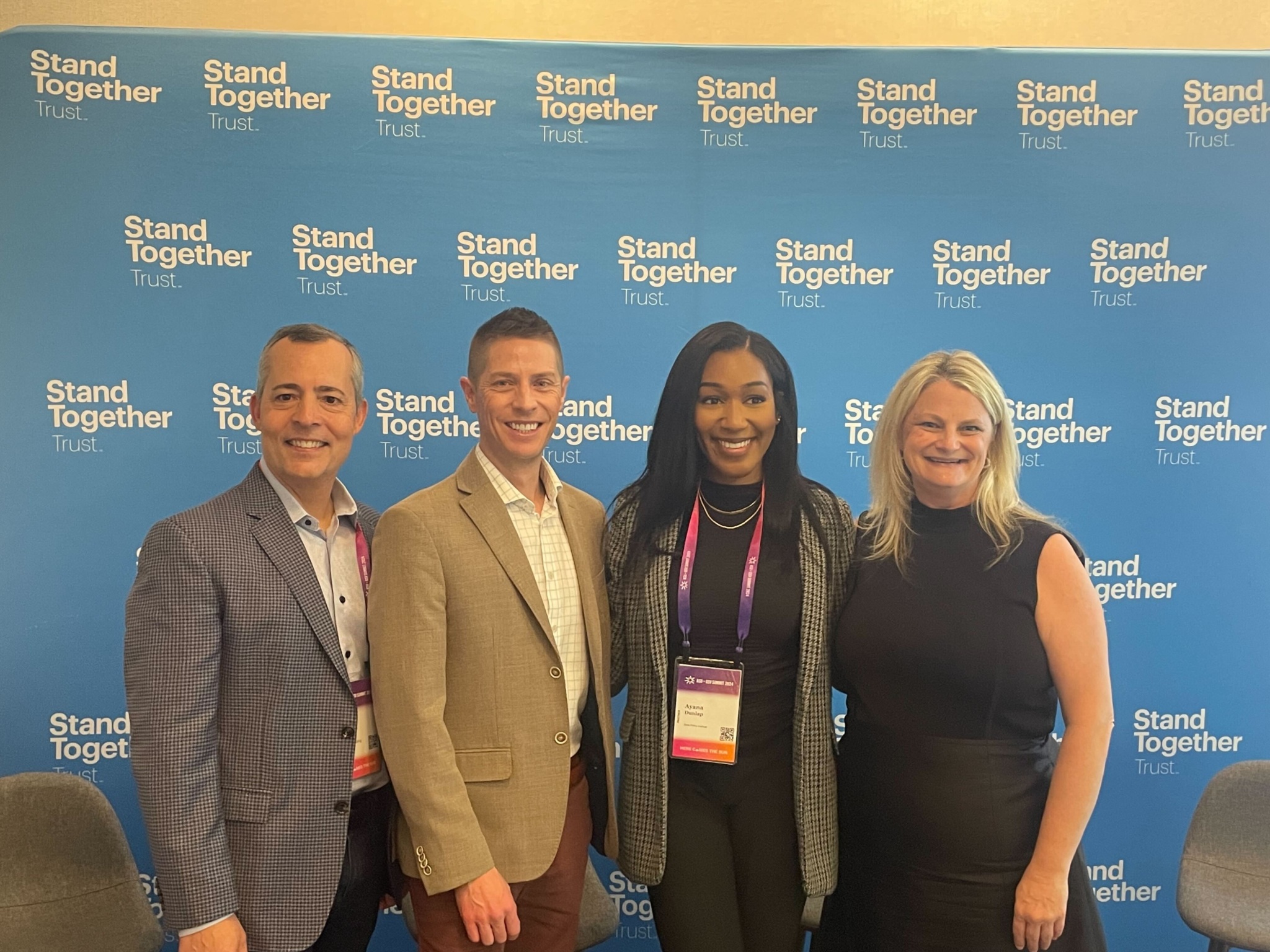 Four smiling adults standing shoulder to shoulder in front of a backdrop that repeats "Stand Together Trust."