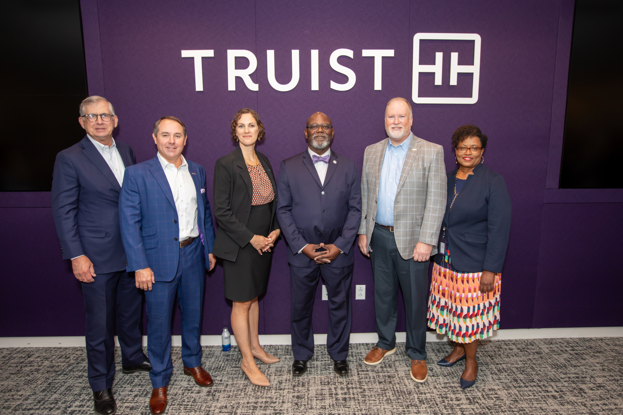 A group of six people in business attire stand in a row smiling in front of a wall with the Truist logo, posing for a group photo.