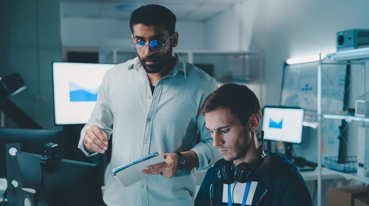 Two colleagues collaborating at a computer workstation, one standing with a tablet and the other seated wearing headphones, focused on a data chart on the screen.