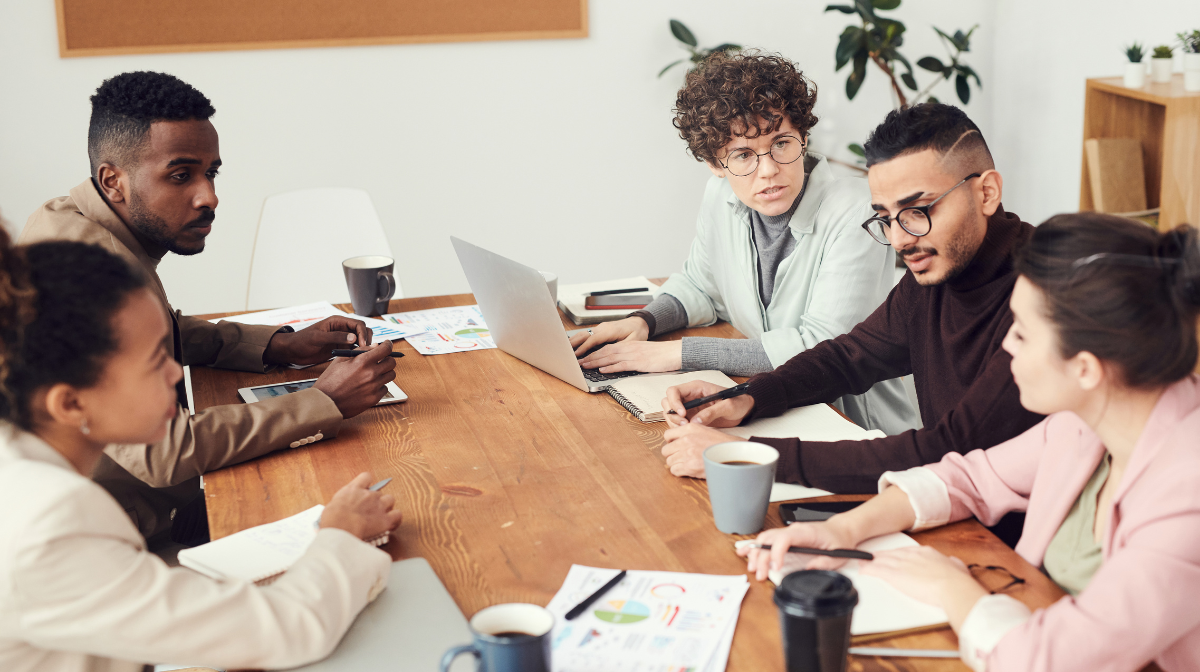 Five colleagues collaborating and conversing around a table, with a laptop, documents, and coffee mugs spread across the table.
