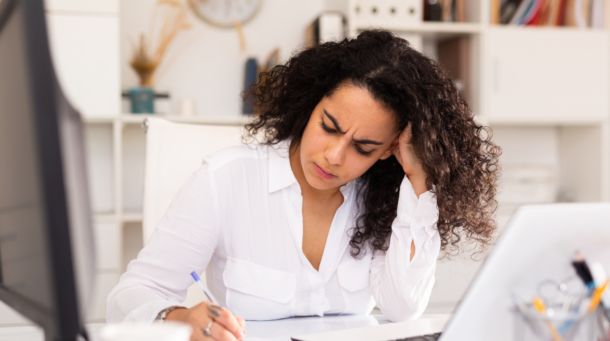 Woman studying at a desk, looking focused while writing notes with a laptop nearby.