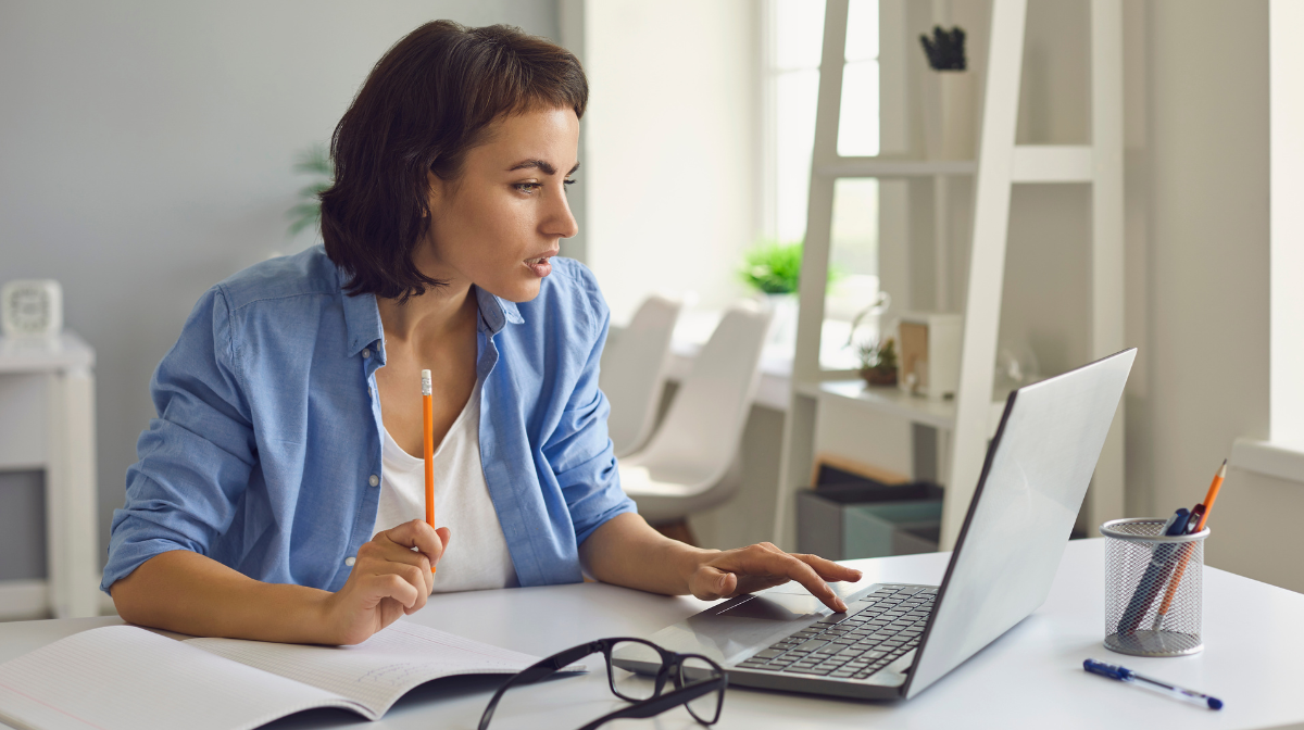 Woman studying at a desk, looking at a laptop screen while holding a pencil and taking notes.