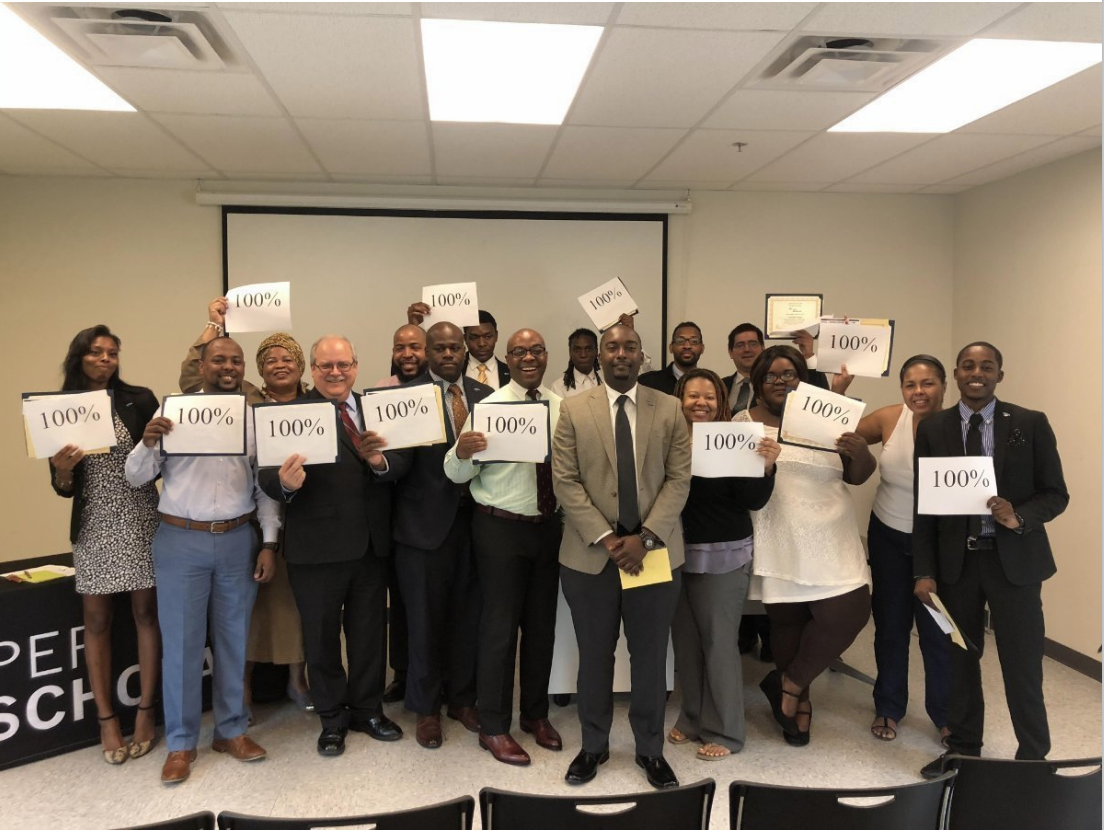 Group of adult graduates standing in a classroom holding signs that read 100%.