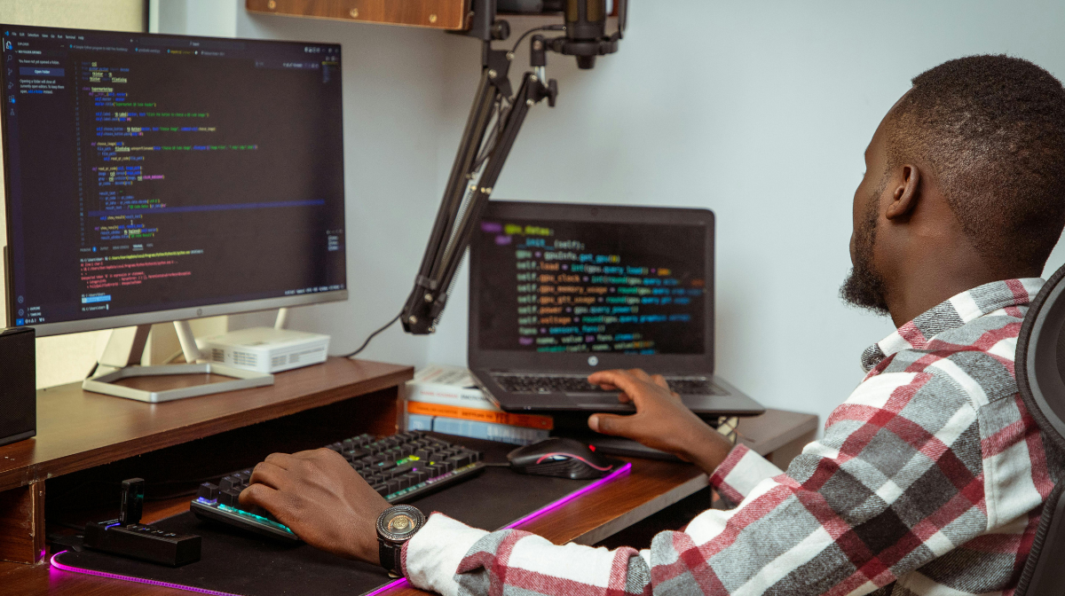 Man working at a desk using a laptop and external monitor displaying code.