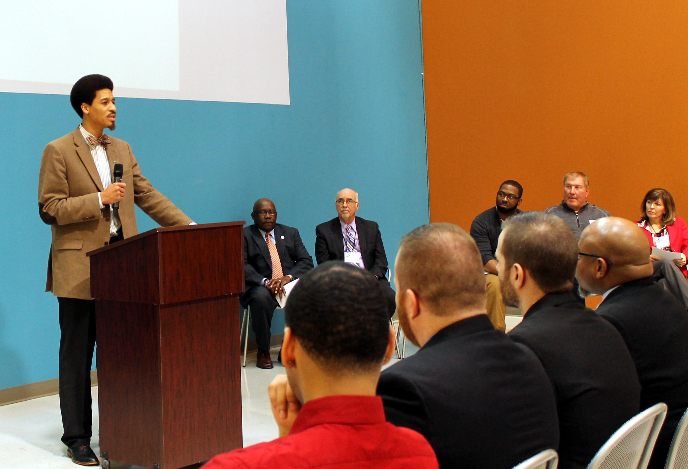 Man standing at a podium speaking to seated audience and panelists during a event.