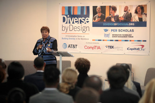 Woman speaking at podium in front of a "Diverse by Design" banner with sponsor logos while an audience listens.