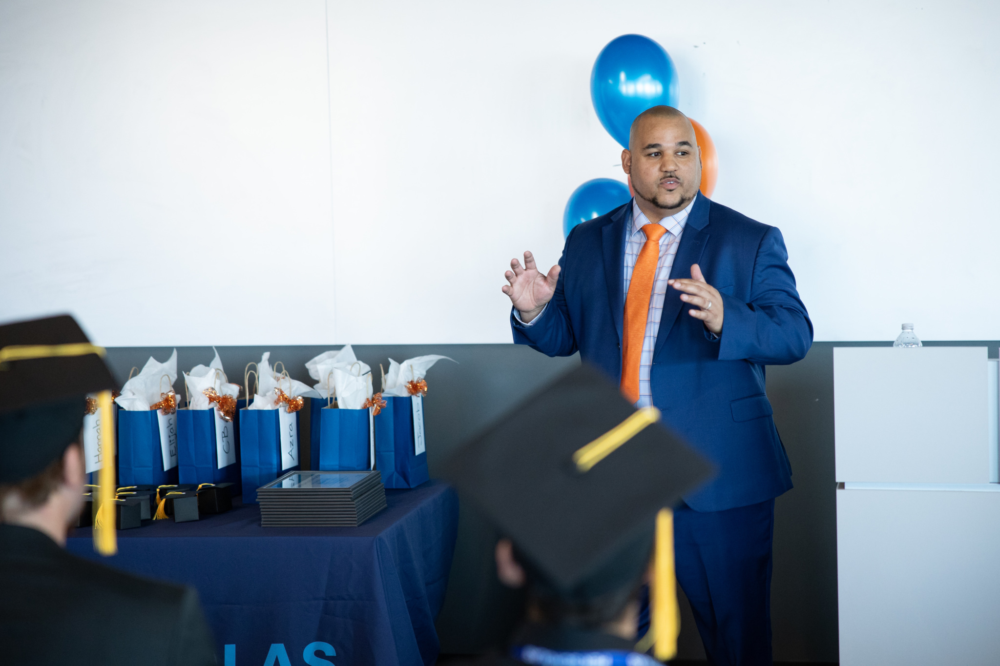 Man in suit and orange tie speaking to a graduation audience beside a table of gift bags and diplomas.