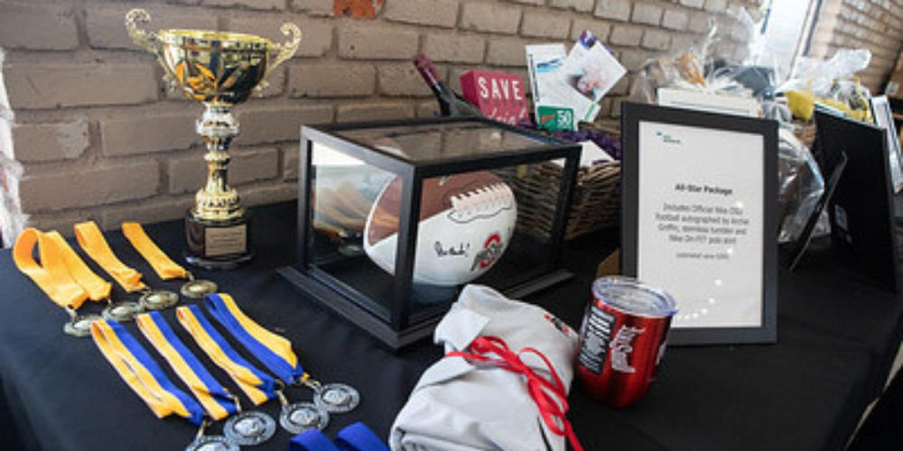 Table of auction items on a table including a trophy, medals, a signed football in a display case, framed bid description, and gift baskets.