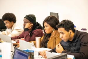 Four adult learners seated at a classroom table working on laptops.