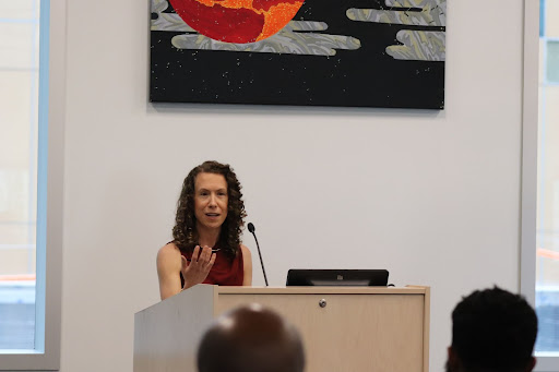 Woman speaking at a podium to an audience, gesturing while presenting.