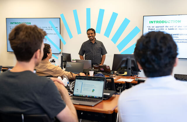 Instructor smiling and speaking to adult students in a classroom with laptops and presentation screens.