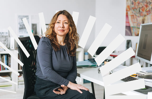 Woman seated at a desk in an office, looking confidently at the camera while holding a phone.