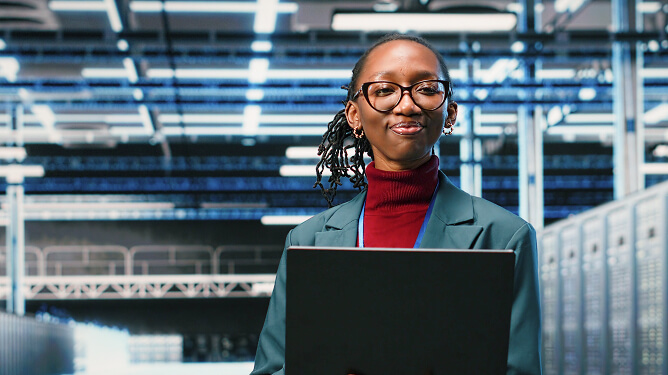 Young female professional confidently standing in a data center holding a laptop.