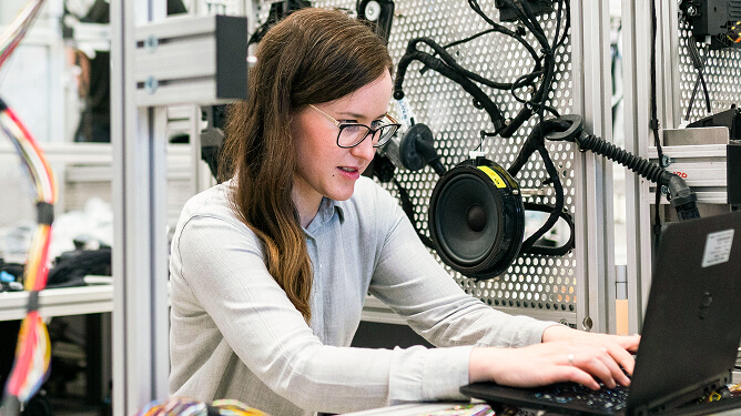 Female trainee working on a laptop next to electronics equipment and speakers in a tech lab.