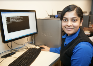 Woman at a computer workstation smiling at the camera while running a program on the monitor.