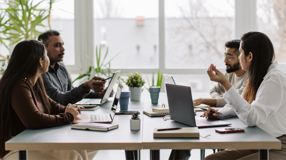 Four adults sit at a table with laptops and notebooks, engaged in a focused discussion in a bright office.
