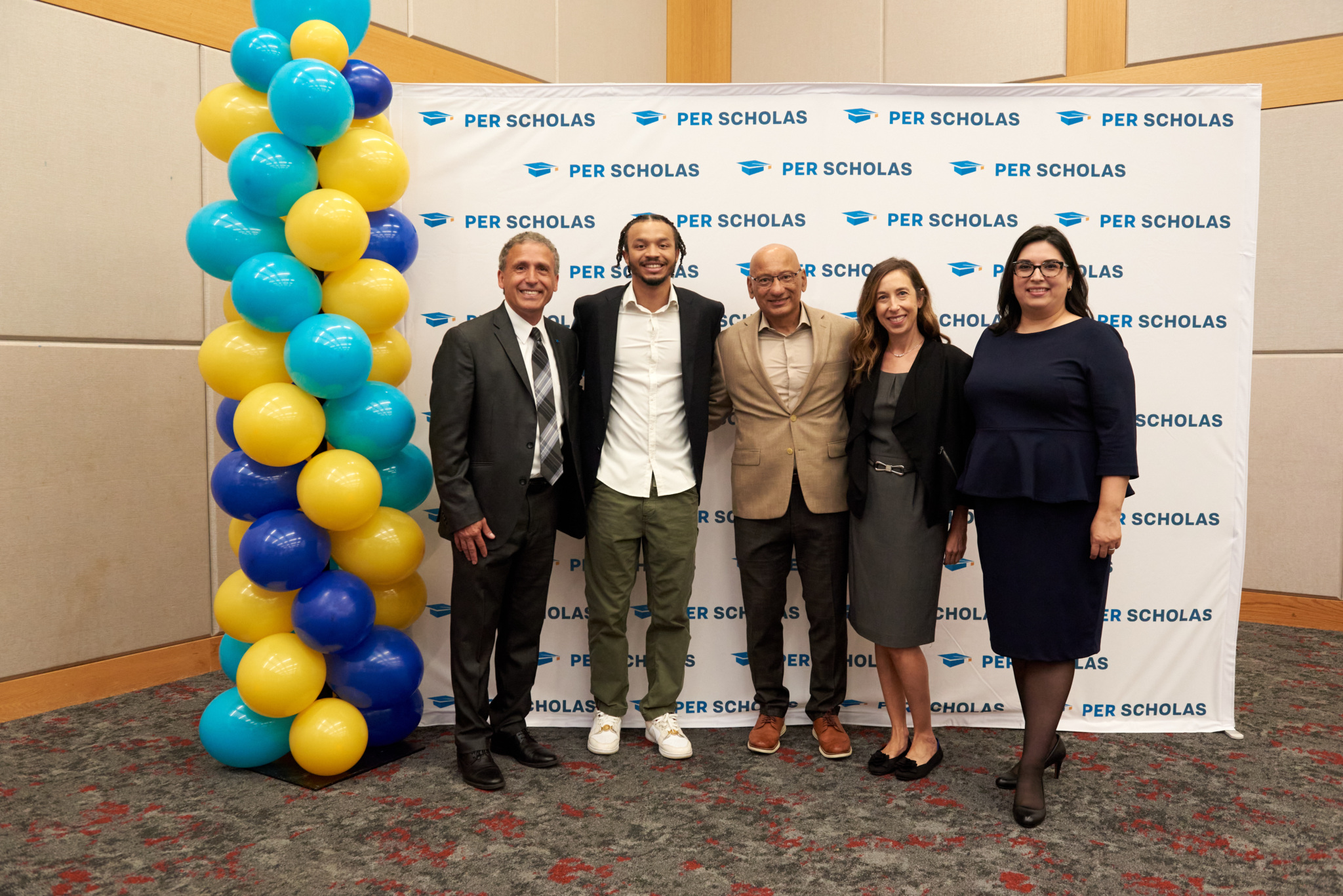 Five smiling adults stand together at a Per Scholas backdrop at an event, posing beside a tall column of blue and yellow balloons.