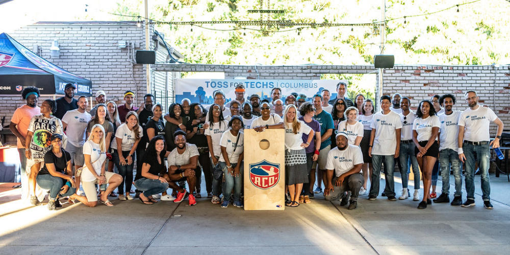 Group of diverse adults standing together at a Toss for Techs Columbus event, posing behind a cornhole board with ACO logo.