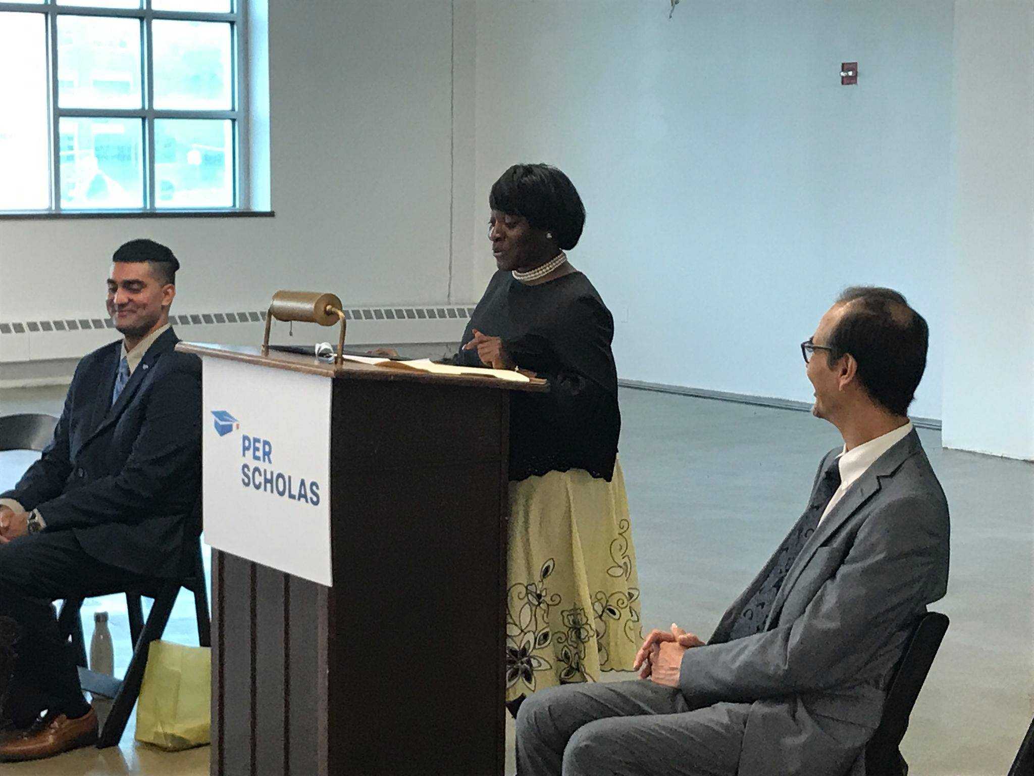 Woman speaking at a podium with a Per Scholas sign while two people sit and listen on each side of her.