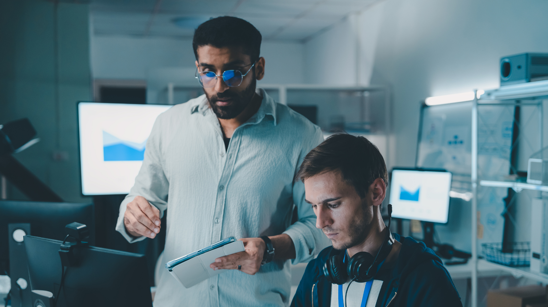 Two colleagues collaborating at a computer workstation, one standing with a tablet and the other seated wearing headphones, focused on a data chart on the screen.