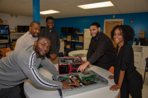 Five adult learners gather around an open desktop computer on a lab table, smiling as they inspect its components.