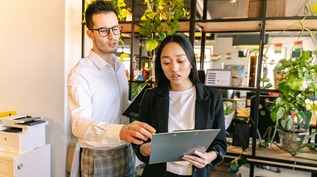 Two coworkers review a clipboard together in an office with plants behind them.