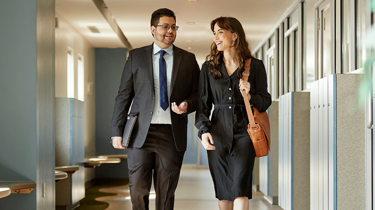 Two professionals walking down an office corridor, engaged in conversation and smiling.