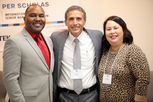 Three smiling adults wearing name badges stand together in front of a Per Scholas banner.
