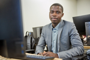 Young man in a classroom setting sits at a computer desk smiling.