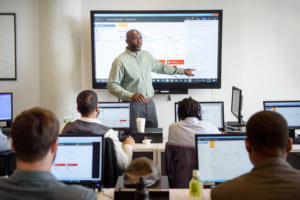 Instructor pointing at large screen while adult students work on computers in a classroom.