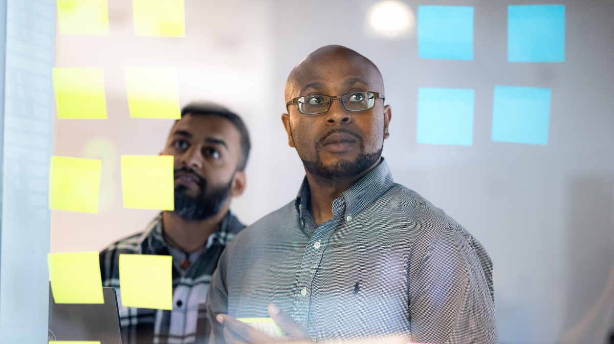 Two colleagues standing behind a glass wall covered in sticky notes, looking thoughtfully at the notes.