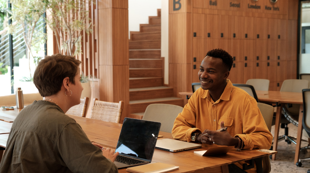 Two people talking and smiling at a wooden table, with a laptop between them in a lounge.