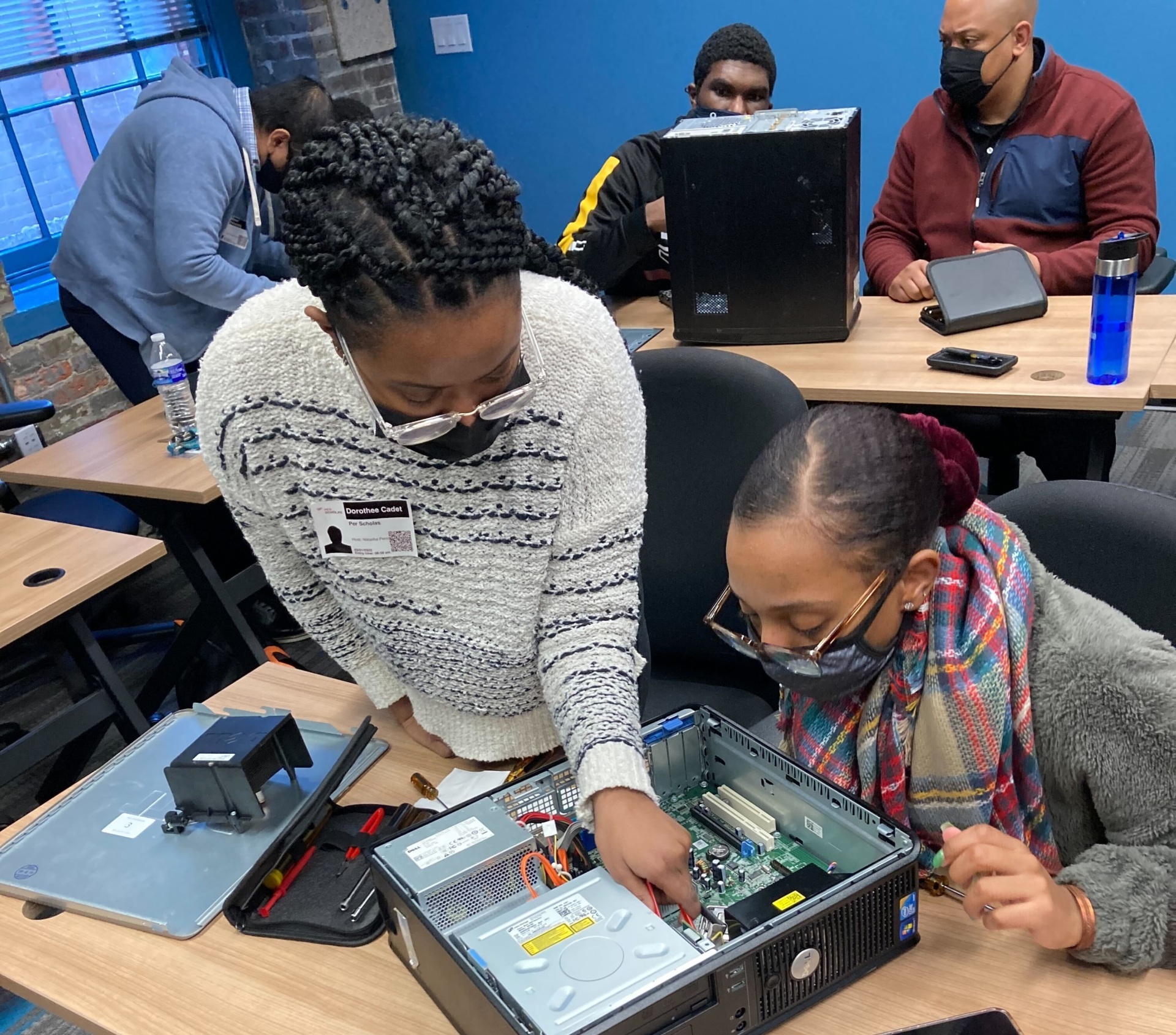Two female learners wearing masks examine and point inside an open desktop computer on a classroom table.