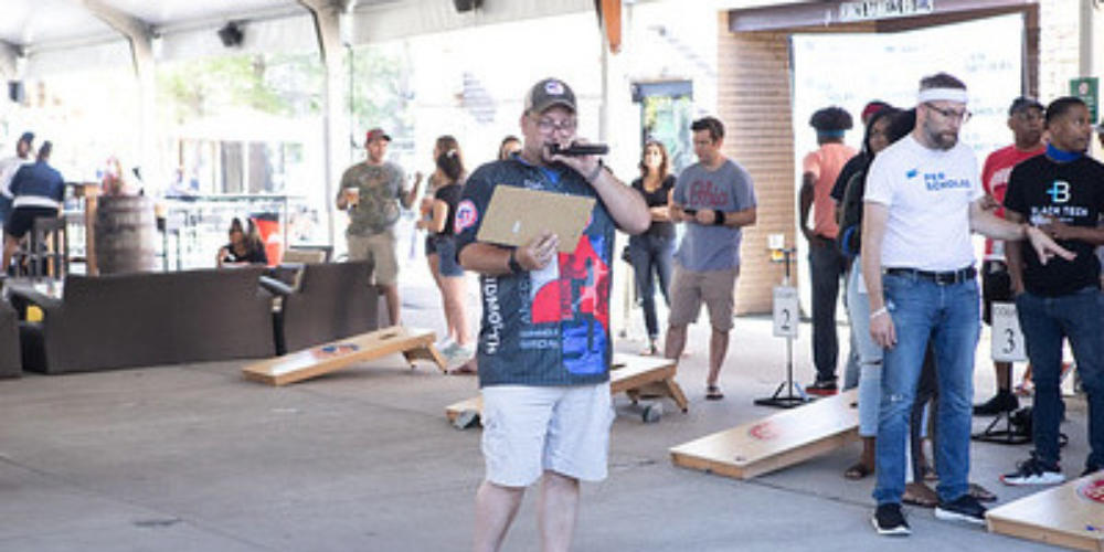 Man holding a clipboard and microphone speaking at a indoor cornhole tournament with players and boards behind him.
