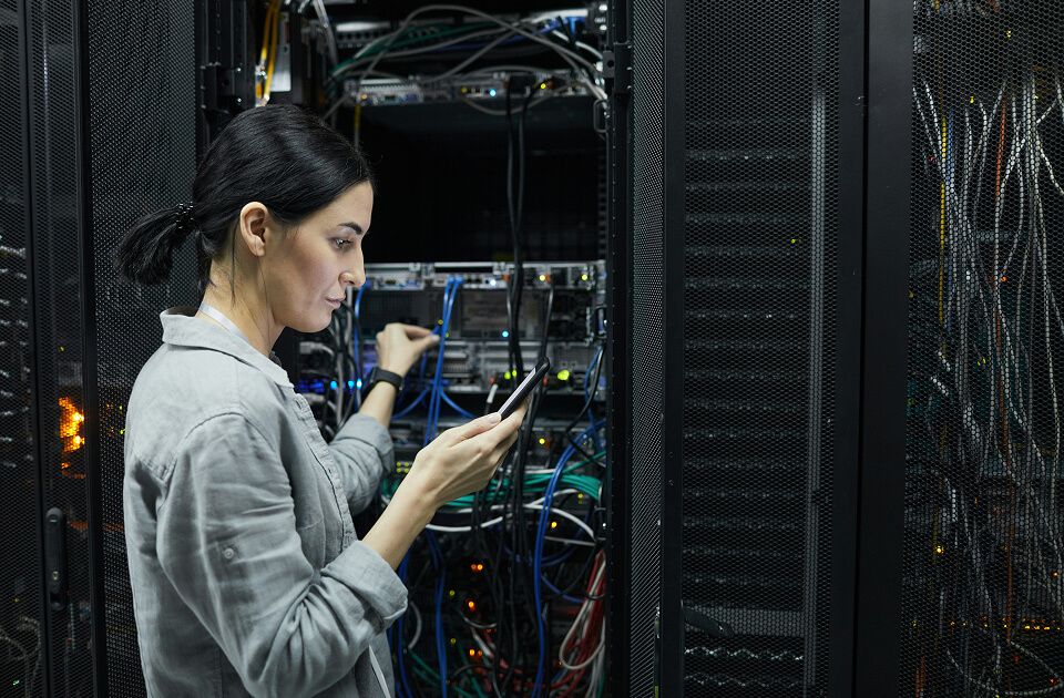 Woman in a gray shirt holding a phone while inspecting cables and equipment in a server rack surrounded by network wiring.