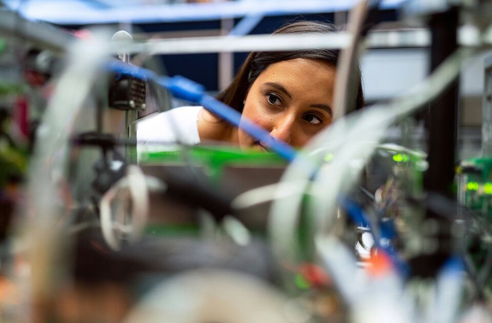 Close-up of a woman with dark hair peering through a tangle of wires and cables while inspecting electrical hardware.
